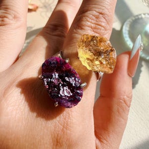 May include: Two rings featuring raw, unpolished gemstones set in silver-toned metal. One ring displays a deep magenta, textured druzy stone, while the other showcases a translucent, golden-yellow citrine crystal with a jagged, natural edge.