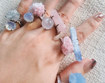 Puede incluir: Una mano que muestra varios anillos de piedras preciosas. Los anillos presentan una variedad de cristales en tonos rosa, azul y transparente, incluyendo cuarzo rosa, cianita y cuarzo. Los anillos tienen engastes plateados.