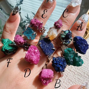 May include: A hand displays several gemstone rings, each with a unique mineral. The rings feature various colors, including pink, blue, green, and yellow. The gemstones are set in silver-toned metal bands, showcasing the natural beauty of the crystals.