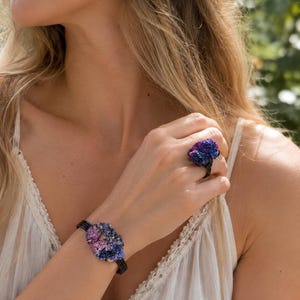 Puede incluir: Un conjunto de joyería compuesto por un anillo y una pulsera, ambos con un racimo de pequeños cristales iridiscentes en tonos azul profundo, morado y rosa. Los cristales están montados sobre finas bandas negras que rodean el dedo y la muñeca.