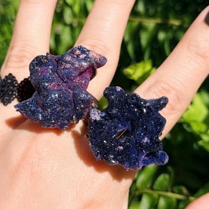 May include: Two rings featuring irregular, textured, dark purple and deep blue clusters with a shimmering, glitter-like finish. The surfaces are uneven and crystalline in appearance, with deep violet and navy tones reflecting light across the jagged, organic shapes.