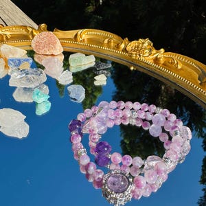 May include: A purple and pink amethyst crystal bracelet with a silver charm. The bracelet is reflected in a mirror with other crystals.