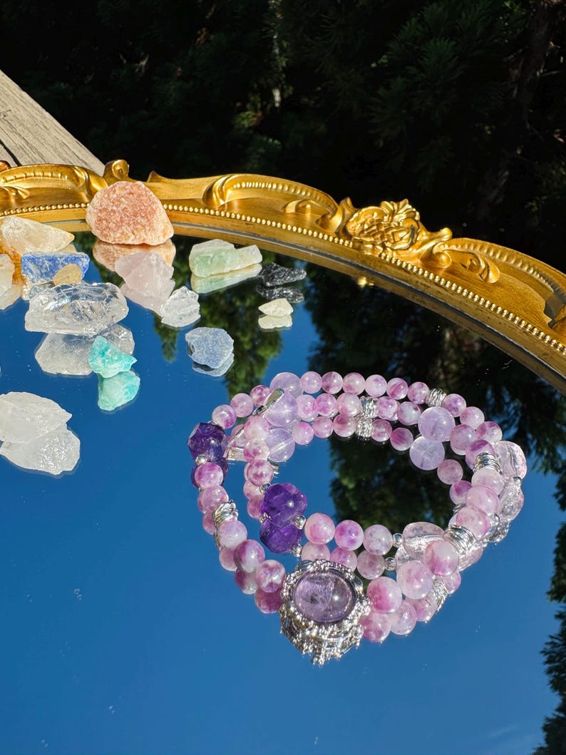 May include: A purple and pink amethyst crystal bracelet with a silver charm. The bracelet is reflected in a mirror with other crystals.