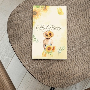 Könnte beinhalten: Ein Hardcover-Tagebuch mit einer Aquarellillustration einer Frau in einem Sonnenblumenhut und -kleid, die Sonnenblumen hält. Das Tagebuchcover trägt die Worte "My Diary" in eleganter Schrift, mit gelben Schmetterlings- und Sonnenblumenakzenten.