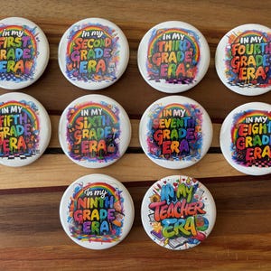 May include: A collection of white round buttons with colorful text and graphics. Each button features the phrase "In My [Grade] Era" in a rainbow-colored, graffiti-style font. The buttons are for grades 1st through 9th, plus a teacher button.