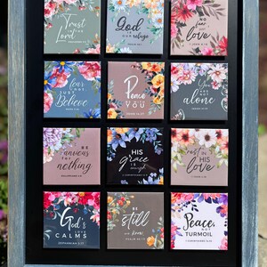 May include: A framed collection of twelve square tiles, each adorned with floral patterns and a religious quote. The tiles are arranged in a grid, showcasing diverse colours and fonts. The frame is a weathered blue-grey.