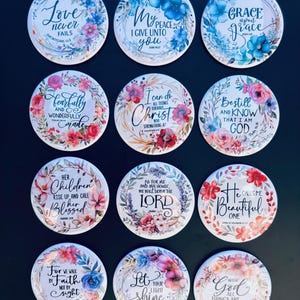 May include: Twelve round coasters with floral designs and inspirational quotes. Each coaster has a different quote in elegant script, surrounded by colourful flowers in shades of pink, blue, and white. The coasters are arranged on a dark surface.