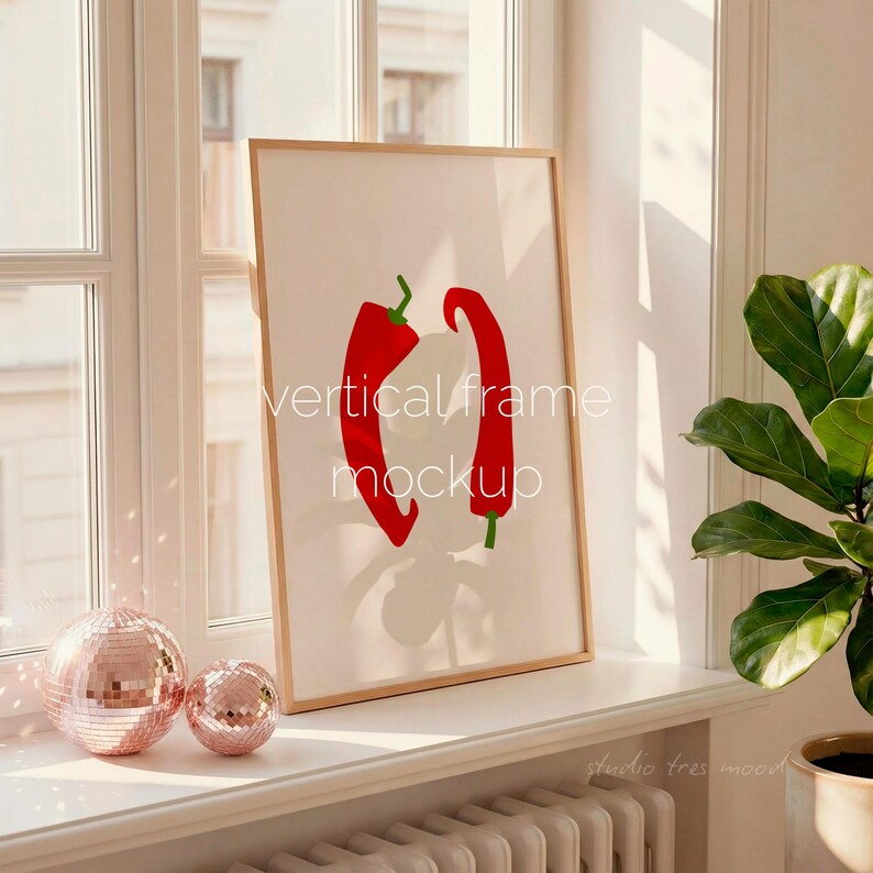 May include: A framed art print featuring a minimalist design of two red chili peppers on a white background. The print is in a light wood frame and is labeled "vertical frame mockup". Two pink disco balls sit on the windowsill.