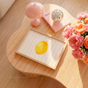 May include: A watercolor lemon print in a wooden frame rests on a light wood table. A pink lamp, a disco ball, and a pink book are also on the table. A bouquet of pink and orange flowers adds a pop of color to the scene.