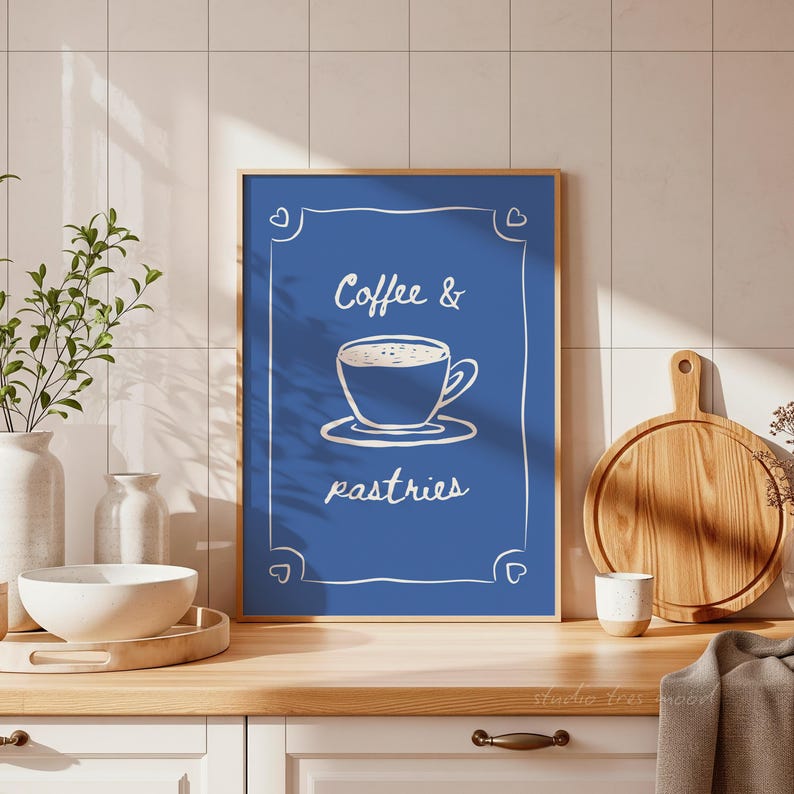 May include: A framed blue print with the words "Coffee & pastries" in white script. The print features a white line drawing of a coffee cup on a saucer. The frame is gold. The print is on a wooden countertop with a white bowl and a wooden cutting board.
