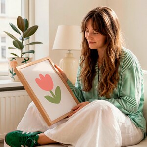 Op de afbeelding: Een ingelijste kunstdruk met een minimalistische illustratie van een roze tulp met een groene stengel en blad. Het kunstwerk staat tegen een witte achtergrond en is ingelijst in natuurlijk hout. De persoon draagt een groen-wit gestreept overhemd.