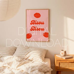 May include: A framed pink art print with the words "Bisou Bisou" in red script, along with three red lip illustrations. The artwork is displayed above a bed with striped bedding and a wooden nightstand.
