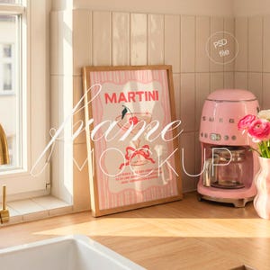 May include: A framed pink and white Martini cocktail print with a cherry garnish. The print is in a light wood frame, displayed on a wooden countertop next to a pink coffee maker and a vase of flowers. The text "Martini" is visible.