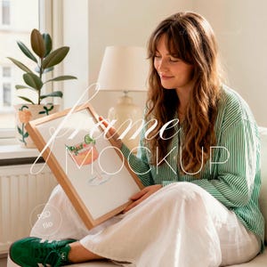 Op de afbeelding: Een ingelijste aquarelprint van een cocktail met een limoenwig, weergegeven in een licht houten frame. De persoon draagt een groen-wit gestreept overhemd en een witte broek. De tekst "frame mockup" staat op de afbeelding.