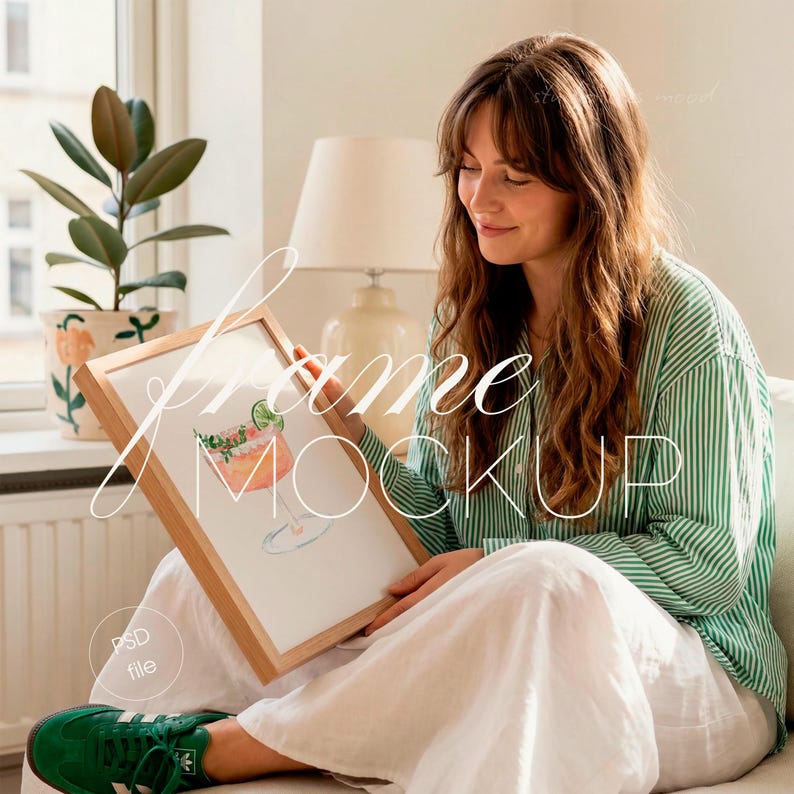 Op de afbeelding: Een ingelijste aquarelprint van een cocktail met een limoenwig, weergegeven in een licht houten frame. De persoon draagt een groen-wit gestreept overhemd en een witte broek. De tekst "frame mockup" staat op de afbeelding.