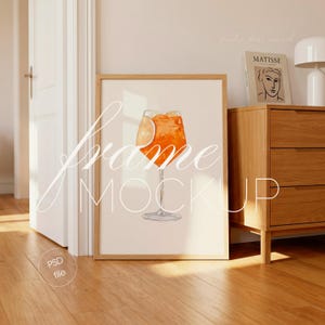 Op de afbeelding: Een ingelijste kunstdruk met een aquarel illustratie van een Aperol Spritz cocktail met een sinaasappelschijfje. Het kunstwerk staat in een licht houten lijst, leunend tegen een muur. De woorden "Frame Mockup" staan op het kunstwerk.