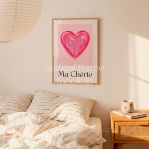 May include: A framed vertical art print featuring a glossy pink heart on a light pink background. The words "vertical frame Ma Ch&eacute;rie mockup" are printed in white below the artwork. The frame is a light wood color.