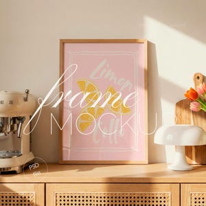 Może przedstawiać: Oprawiony różowy nadruk z motywem plasterków cytryny i napisem "Lemon Calls". Nadruk jest w jasnej drewnianej ramie, eksponowany na drewnianej szafce. Widoczna jest również biała lampa i ekspres do kawy.