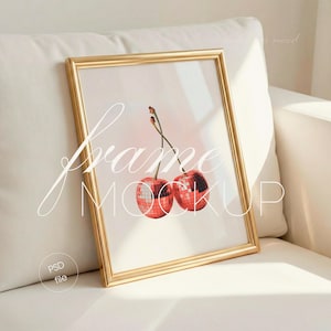 May include: A gold rectangular picture frame with a print of two red cherries with disco ball textures. The frame is leaning against a white cushion. The words "frame mockup" are written on the print.