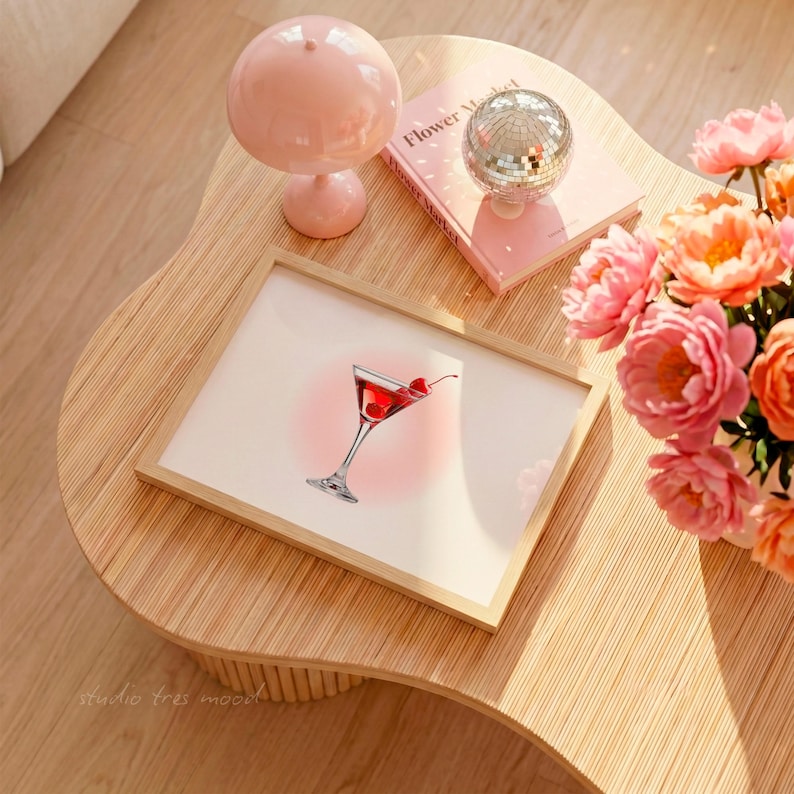 May include: A framed print of a cocktail with cherries, set on a light wood table. A pink lamp, a disco ball, and a book titled "Flower Market" are also on the table. A bouquet of pink flowers adds a pop of color to the scene.
