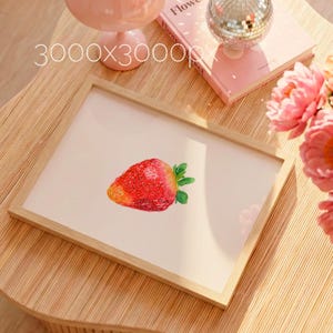 May include: A framed print featuring a watercolor illustration of a ripe strawberry. The strawberry is red with yellow and green accents, set against a light pink background. The frame is made of light-colored wood. The image is on a wooden table.