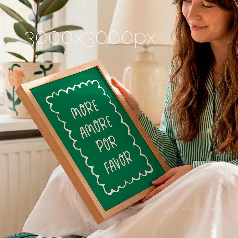 Op de afbeelding: Een ingelijst groen schoolbord met de woorden "MORE AMORE POR FAVOR" in witte letters. Het frame is gemaakt van lichtgekleurd hout. De afbeelding wordt vastgehouden door een persoon die een groen-wit gestreept overhemd draagt.
