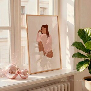 May include: A framed art print featuring a minimalist illustration of a person in a pink sweater. The artwork is displayed on a windowsill, alongside two rose-gold disco balls. The print has a light brown wooden frame. The text "studio tres mood" is visible on the windowsill.