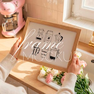 Könnte beinhalten: Ein hellbrauner Holzrahmen-Mockup, der von einer Person gehalten wird und eine schwarz-weiße Illustration von Kaffeezubereitungsgeräten zeigt. Der Rahmen wird in der Nähe eines Waschbeckens mit Blumen und einem rosa Küchenmixer im Hintergrund gehalten.
