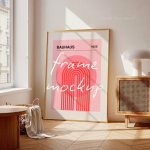 Op de afbeelding: Een gouden ingelijste poster mockup tegen een muur. De poster heeft een roze en rood ontwerp met het woord "BAUHAUS" en het jaar "1925". De woorden "frame mockup" staan op de poster. De kamer heeft houten vloeren en een bijzettafel.