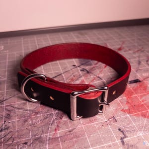 May include: A black leather collar with a vibrant red interior lining. The collar features a polished silver-tone metal buckle and a matching D-ring for attachment. The design includes multiple adjustment holes along the strap for a secure fit.