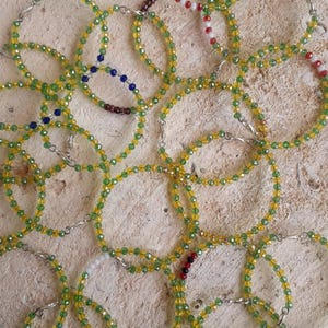 May include: A selection of beaded bracelets in yellow and green hues, with red, white, and blue bead accents. The bracelets are linked with silver-coloured clasps, creating a layered effect. The bracelets are displayed on a light brown surface.