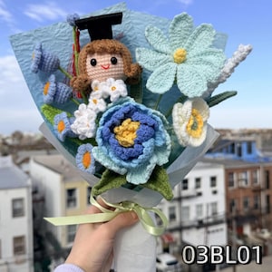 May include: A crochet bouquet featuring a doll wearing a black graduation cap with a red tassel, surrounded by blue, white, and light blue crocheted flowers and green leaves. The arrangement is wrapped in light blue paper with a light green ribbon.