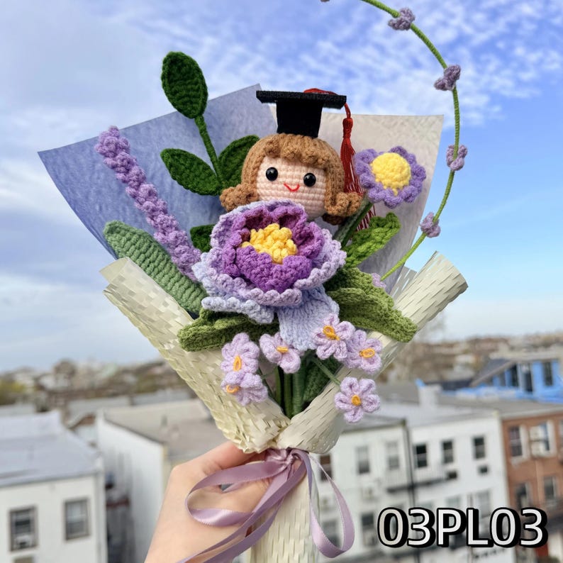 May include: A crochet bouquet featuring a doll wearing a black graduation cap, a large purple flower with a yellow center, lavender sprigs, and small light purple flowers with yellow centers, all wrapped in textured paper with a purple ribbon.