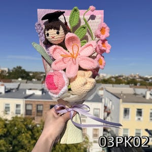 May include: A crochet bouquet featuring a small doll wearing a black graduation cap, a large pink lily, a pink and white rose, a peach-colored flower, and green leaves. The arrangement is wrapped in pink paper and tied with a light purple ribbon.