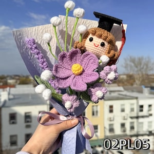 May include: A crochet bouquet featuring a doll wearing a black graduation cap with a red tassel, a large purple flower with a yellow center, smaller purple flowers, and white spherical buds on green stems, all wrapped in light purple paper with a ribbon.
