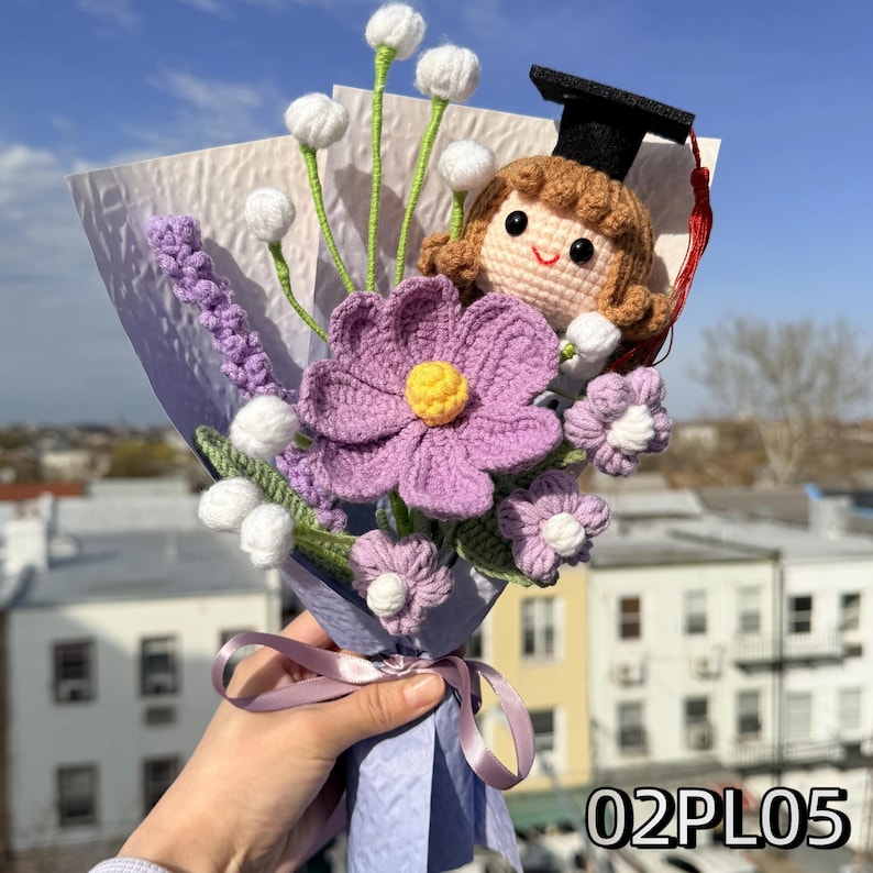 May include: A crochet bouquet featuring a doll wearing a black graduation cap with a red tassel, a large purple flower with a yellow center, smaller purple flowers, and white spherical buds on green stems, all wrapped in light purple paper with a ribbon.