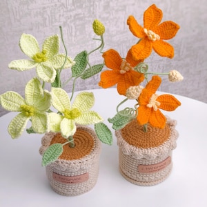 May include: Two crochet flower arrangements in small pots. One arrangement features light green flowers, the other orange. Both have green leaves and are in beige pots with "HANDMADE" labels. The flowers are on a white surface.