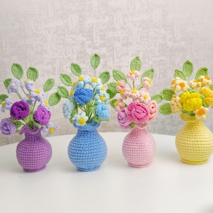 Könnte beinhalten: Vier gehäkelte Blumenarrangements in kleinen Vasen. Die Vasen sind in Lila, Blau, Rosa und Gelb gehalten. Die Blumen sind in Lila-, Blau-, Rosa- und Gelbtönen gehalten, mit grünen Blättern. Die Arrangements stehen auf einer weißen Oberfläche.