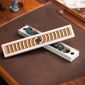 May include: A white rectangular dice box with a sliding lid featuring a black, orange, and white striped pattern. The box is open, revealing blue translucent dice inside. The word Wyrmwood is embossed on the side of the white container.
