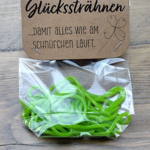 Könnte beinhalten: Ein klarer Plastikbeutel gefüllt mit hellgrünen Gummibändern. Oberhalb des Beutels befindet sich ein braunes Papieretikett mit den Worten "Glückssträhnen" und "...damit alles wie am schnürchen läuft." in schwarzer Schrift, sowie eine Kleeblatt-Illustration.
