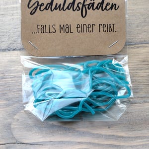 Könnte beinhalten: Eine transparente Tüte mit teal Gummibändern, darüber eine braune Karte. Auf der Karte stehen die Worte "Geduldsfäden" und "...FALLS MAL einer reißt." in Schwarz.