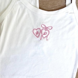 Op de afbeelding: Witte tanktop met roze geborduurde harten. Eén hart heeft de letter "B" en de andere de letter "D" met een strik. De tanktop heeft een ronde hals en dunne bandjes. Het woord "Jones" is blauw op de top geborduurd.