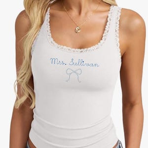 Puede incluir: Camiseta sin mangas blanca con un ribete de encaje alrededor del escote. El texto "Mrs. Sullivan" está escrito en azul, con un lazo azul debajo. La camiseta sin mangas es acanalada.
