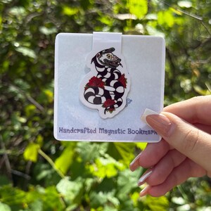 May include: A white magnetic bookmark with a black and white illustration of a snake with red roses. The bookmark has the text "Handcrafted Magnetic Bookmark" printed on the bottom.