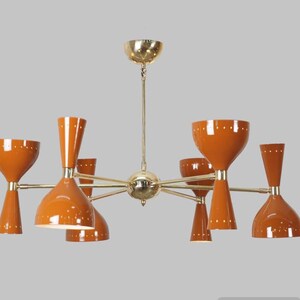 Op de afbeelding: Een mid-century moderne messing en oranje kroonluchter met zes lichten. De kroonluchter heeft een stervormig ontwerp met armen die zich uitstrekken vanaf een centrale naaf. Elke arm heeft een armatuur met een klokvormige kap.