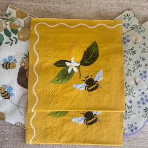 May include: A collection of decorative paper napkins with bee and floral designs. The napkins feature a variety of patterns, including a yellow napkin with a bee and flower illustration, and napkins with floral and honeycomb patterns.