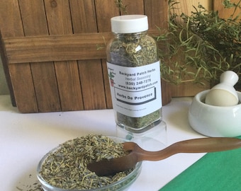 May include: A glass jar filled with dried herbs labeled "Backyard Patch Herbs Herbal Seasoning (830) 248-7278 www.backyardpatch.com Herbs De Provence". The jar is sitting on a wooden surface next to a white mortar and pestle. A small bowl of dried herbs and a wooden spoon are in the foreground.