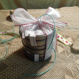 May include: A stack of four round metal tins with a white ribbon tied around them. The tins are decorated with a white and red polka dot ribbon and a white label with the text "www.cozyjam.com".