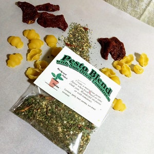 May include: A clear plastic bag filled with a green pesto blend with sun-dried tomatoes. The bag has a white label with the text "Pesto Blend with Sun-Dried Tomatoes" and a description of the ingredients. The label also includes a small illustration of a potted herb plant.