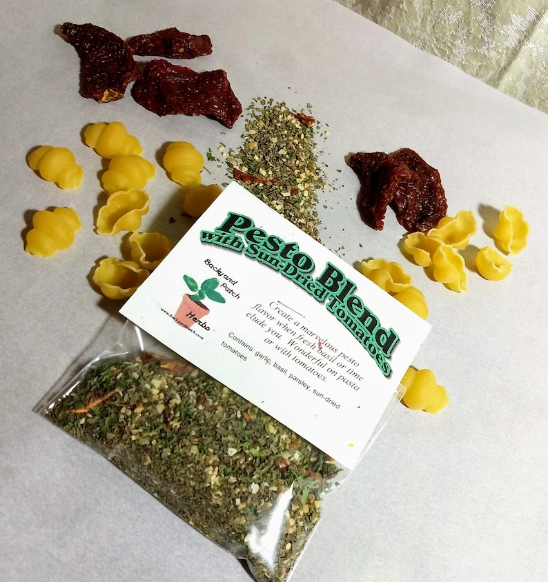 May include: A clear plastic bag filled with a green pesto blend with sun-dried tomatoes. The bag has a white label with the text "Pesto Blend with Sun-Dried Tomatoes" and a description of the ingredients. The label also includes a small illustration of a potted herb plant.